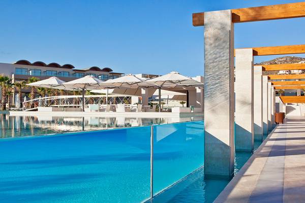 Außenbereich des Avra Imperial Hotels mit Pool, weißen Sonnenschirmen und Betonsäulen.