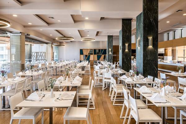 Innenansicht des Restaurants im Avra Imperial Hotel mit vielen weißen Tischen und Stühlen, die für Gäste vorbereitet sind.