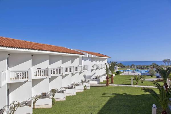 Außenansicht des Mrs Chryssana Beach Hotels mit weißen Gebäuden und roten Dächern, umgeben von grüner Rasenfläche und Palmen.