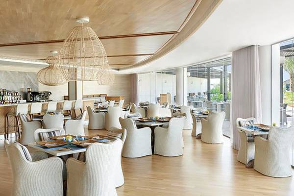 Innenansicht des Restaurants im Waldorf Astoria Ras Al Khaimah mit weißen Stühlen und Tischen, Holzboden und großen Glasfenstern.