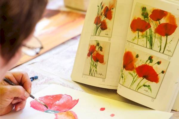 Eine Person malt eine rote Blume auf einem Blatt Papier, während ein offenes Buch mit Blumenbildern danebenliegt.