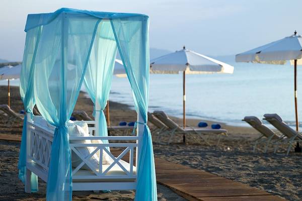 Strandszene mit einem weißen Bett unter blauen Vorhängen und Sonnenschirmen am Meer.