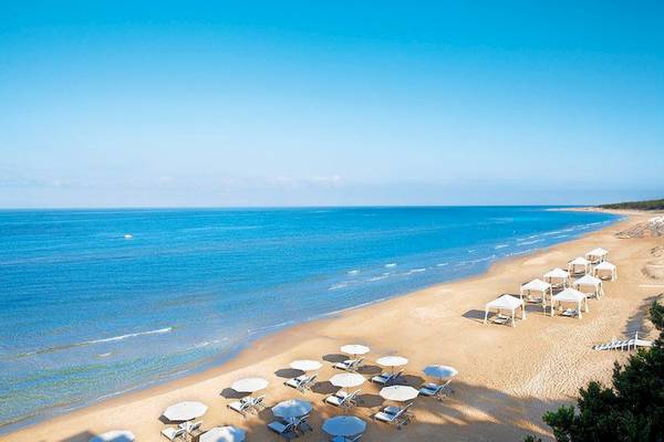 Luftaufnahme des Strandes des Grecotel La Riviera & Aqua Park Hotels mit weißen Sonnenschirmen und Liegen.