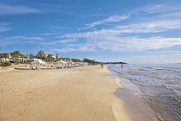 Strand des Grecotel La Riviera & Aqua Park mit weißen Sonnenschirmen und Hotels im Hintergrund.