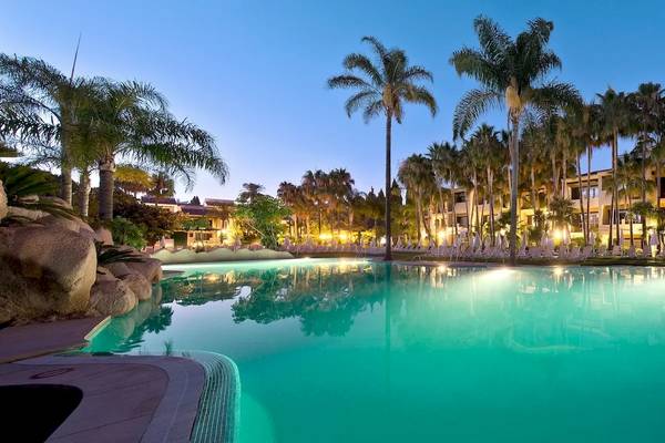 Abendansicht des Poolbereichs des Bluebay Banus Hotels mit leuchtenden Palmen und gesäumten Sonnenliegen.
