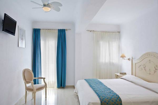 Schlafzimmer im Kamari Hotel mit weißem Bett, blauen Vorhängen und Wand-TV.