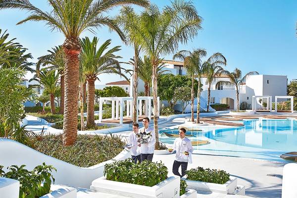 Drei Männer im weißen Outfit stehen vor dem Grecotel Caramel Boutique Resort und präsentieren eine Flasche Wein.