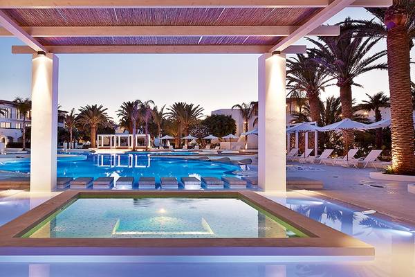 Der Außenpool des Grecotel Caramel Boutique Resort ist bei Einbruch der Dunkelheit beleuchtet und zeigt die umgebenden Gebäude und Palmen.