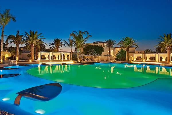 Nachtansicht des Swimmingpools im Grecotel Caramel Boutique Resort mit farbigen Lichtern und Palmen.