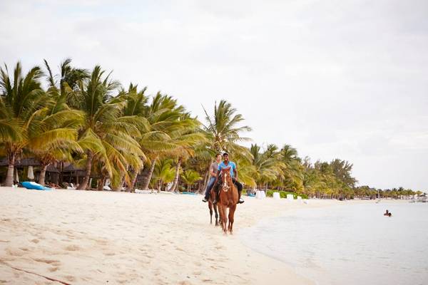 Zwei Personen reiten auf Pferden entlang eines Strandes des LUX Le Morne Hotels.