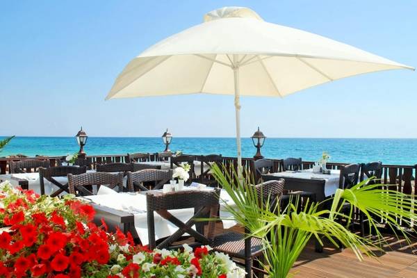 Terrasse des Riviera Beach Hotels mit Blick auf das Meer und Tischen mit Sonnenschirmen und Blumen.