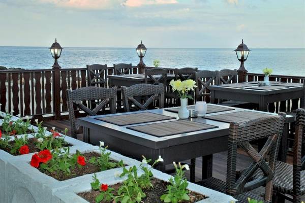 Terrasse des Riviera Beach Hotels mit Blick auf das Meer und mehreren Tischen und Stühlen, die von Blumenbeeten umgeben sind.