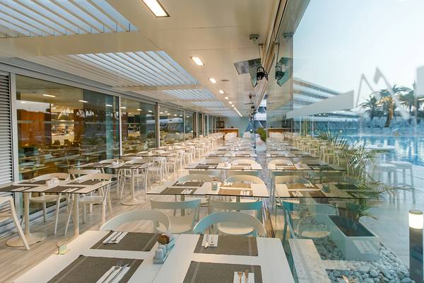 Innenansicht des Restaurants im Santa Monica Suites Hotel mit vielen Tischen und Stühlen, großen Glaswänden und einer Poolreflexion.