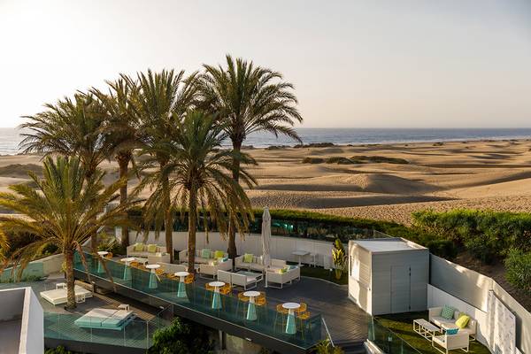 Terrasse des Santa Monica Suites Hotels mit Palmen, weißen Möbeln und Blick auf das Meer und die Dünen.
