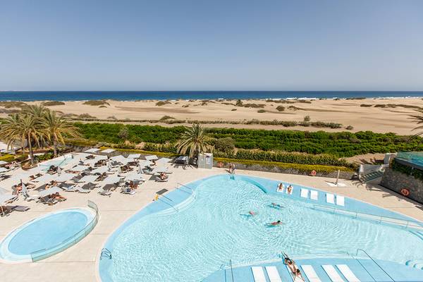 Ein Hotelpoolbereich mit Blick auf eine sandige Küste und mehreren Schwimmern.