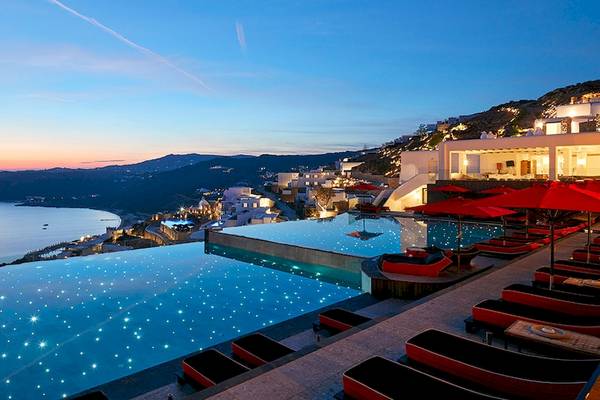 Abendlicher Ausblick auf das Myconian Avaton Resort mit Infinity-Pool und roten Sonnenschirmen.