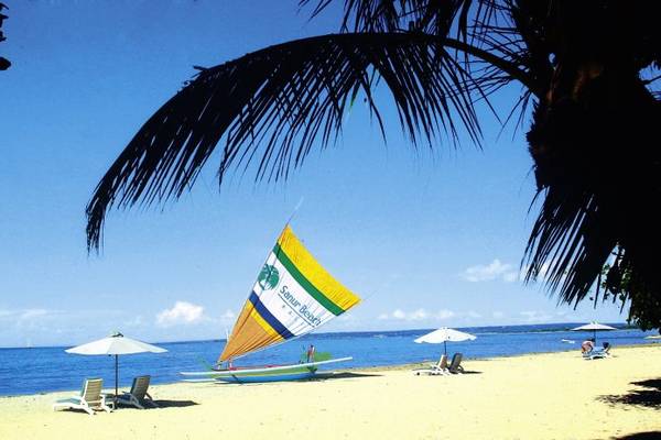 Strandszene des Prama Sanur Beach Bali Hotels mit Segelboot, Sonnenschirmen und Sonnenliegen