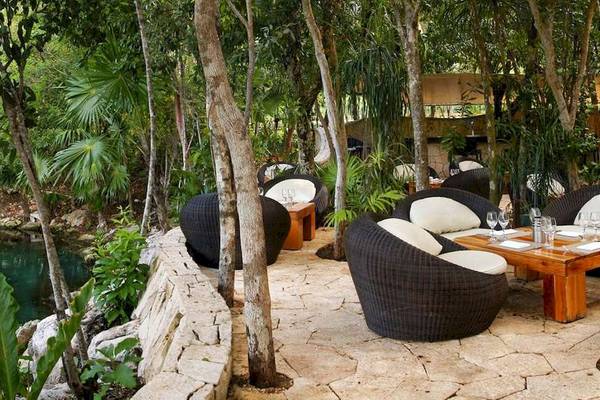Gartensitzplatz im Catalonia Royal Tulum mit Tischen, Stühlen und Blick auf einen kleinen Fluss.