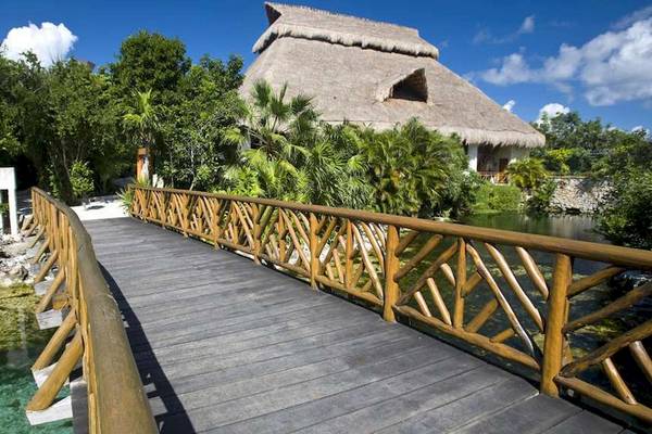 Holzbrücke zum Hotel Catalonia Royal Tulum mit Blick auf ein traditionelles Dach und umgebende Pflanzen