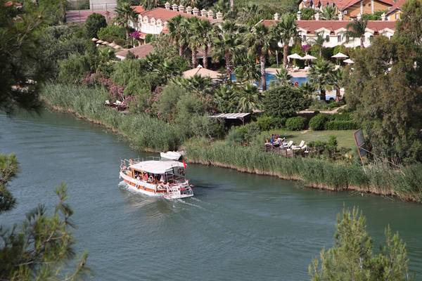Boot auf dem Fluss vor dem Dalyan Resort Hotel mit umliegenden Gebäuden und Palmen.