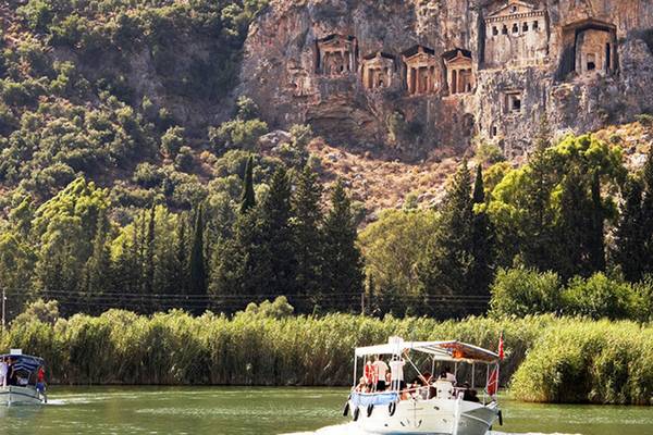 Zwei Boote fahren auf dem Dalyan Fluss vor Felsengräbern.