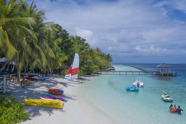 Strandansicht des Royal Island Resort & Spa mit Palmen, Booten und Schwimmern im Wasser.