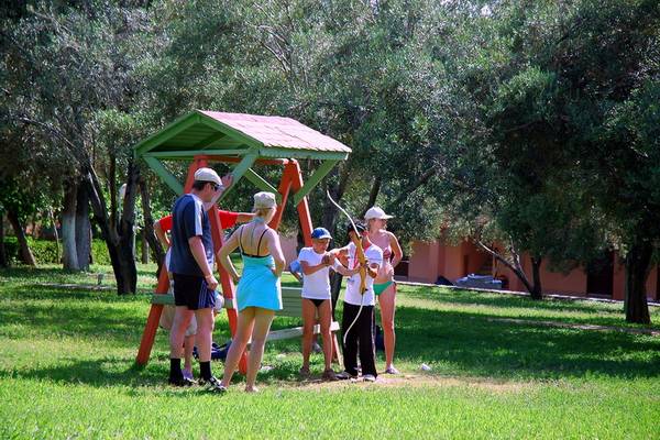 Gruppe von Kindern und Erwachsenen übt Bogenschießen in einem Park, hinter ihnen ein buntes Gebäude.
