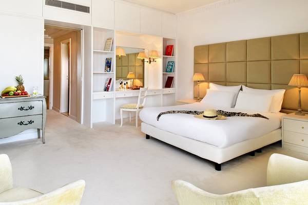 Innenansicht eines Schlafzimmers im Mitsis Grand Hotel Beach mit weißem Bett, Schreibtisch und Früchten auf dem Nachttisch.