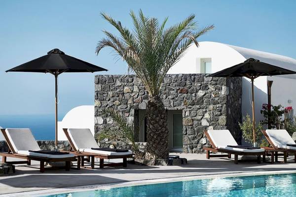 Außenbereich des Santo Pure Oia Suites mit Loungestühlen, Palme und Sonnenschirmen neben dem Pool.