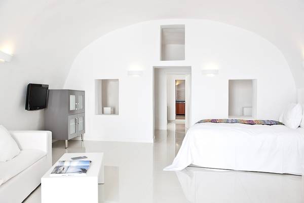 Innenansicht eines Schlafzimmers im Katikies Chromata Santorini Hotel mit weißem Bett, Couch, Tisch und Fernseher.
