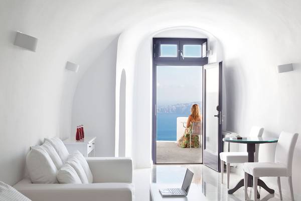 Ein helles Zimmer mit offenem Balkon und Meerblick, Möbel sind weiß und die Frau sitzt draußen.