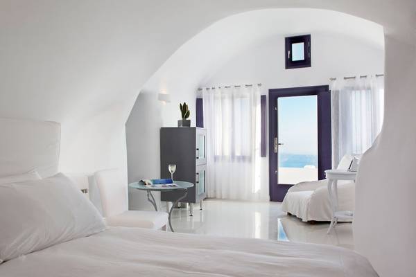 Schlafzimmer des Katikies Chromata Santorini Hotels mit weißem Bett, Tisch und Dekor.