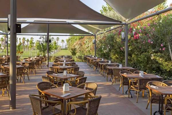 Terrasse des Royal Seginus Hotels mit vielen runden Tischen und Stühlen unter Sonnensegeln.