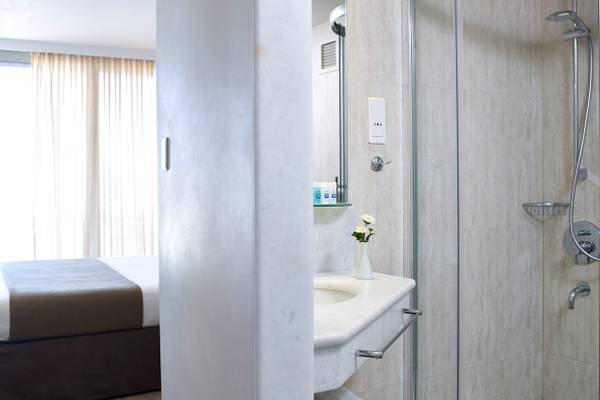 Badezimmer im Kriti Beach Hotel mit Dusche, Waschbecken und Bett im Hintergrund.