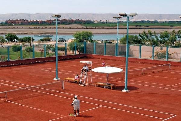 Tennisplatz im Movenpick Resort & Spa El Gouna mit Spielern, einer Tribüne und einem See im Hintergrund.