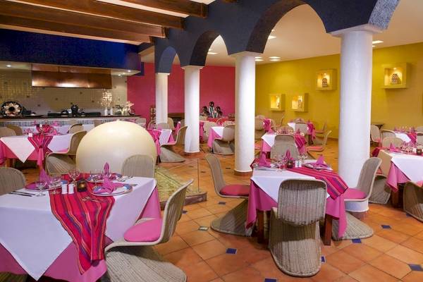 Innenansicht des Restaurants im Grand Sirenis Riviera Maya Resort & Spa mit pinken und gelben Wänden, runden Tischen und Stühlen.