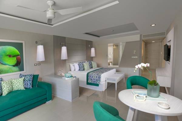 Innenansicht eines Zimmers im Grand Sirenis Riviera Maya Resort & Spa mit grünem Sofa, Bett, Tisch und Kunstwerk an der Wand.