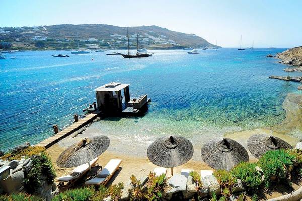 Kivotos Mykonos Strand mit Booten im Wasser und Sonnenschirmen auf der Wiese.