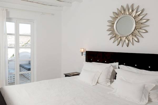Schlafzimmer im Kivotos Mykonos Hotel mit weißem Bett, Kopfteil und weißen Kissen, Spiegel und Lampe an der Wand, und Fenster mit Vorhängen.