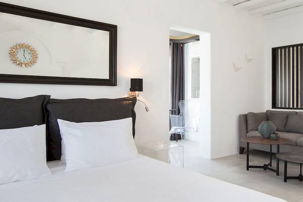 Ein modernes Hotelzimmer von Kivotos Mykonos mit weißem Bett, grauem Sofa, Tisch und Uhrspiegel.