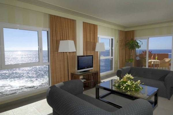 Wohnzimmer des Roca Nivaria Gran Hotel mit Meerblick, zwei Sofas, Tisch mit Blumen, Fernseher und großen Fenstern.