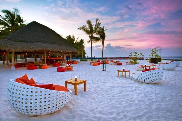 Strandloungebereich des Kuramathi Maldives Hotels bei Sonnenuntergang mit Strohhütten, roten Kissen und Laternen.