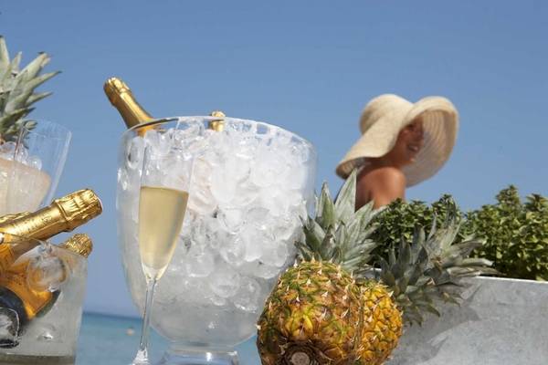 Eine Flasche Champagner und Gläser auf Eis in einem Glasgefäß, umgeben von Ananas und Pflanzen, mit einer Person im Hintergrund.