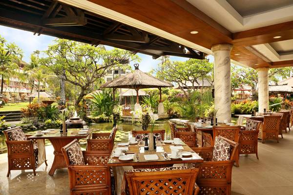 Innenansicht des Restaurants im Nusa Dua Beach Hotel & Spa mit runden Tischen und Stühlen, umgeben von Pflanzen und einem Wasserbrunnen.