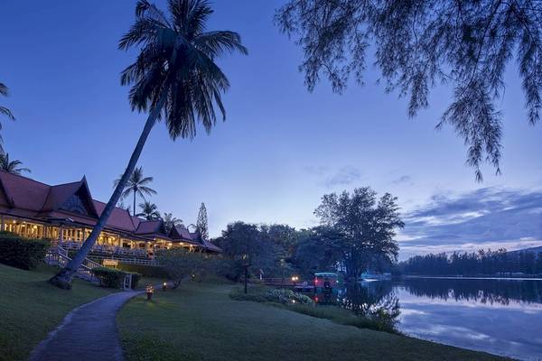 Außenansicht des Dusit Thani Laguna Phuket Hotels bei Abendbeleuchtung mit Palmen und Wasser.