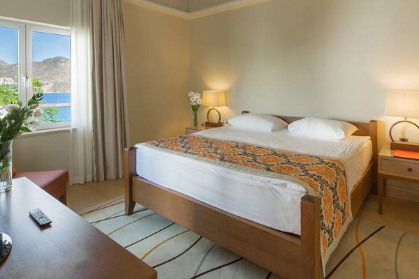 Hotelzimmer im Xanadu Island Bodrum mit Blick auf das Meer und Bergen, ein Doppelbett mit weißen Kissen und orangefarbenem Bettdeckenbezug.