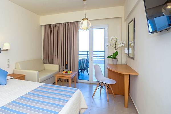 Innenansicht eines Hotelzimmers im Themis Beach Hotel mit Bett, Schreibtisch und Balkon.