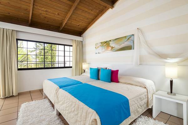 Schlafzimmer im BlueBay Villas Doradas mit zwei Betten, blauen Decken, bunten Kissen und einem Fenster mit Aussicht auf Bäume.