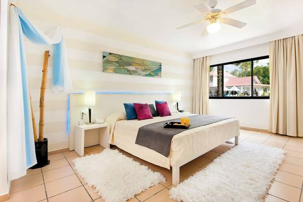Schlafzimmer im BlueBay Villas Doradas mit einem Bett, zwei Nachttischen, Deckenventilator und einem Fenster mit Blick auf ein Gebäude und Bäume.