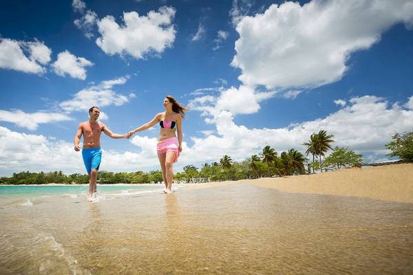 Ein Paar läuft Hand in Hand auf einem Strand mit blauem Himmel und Wolken.